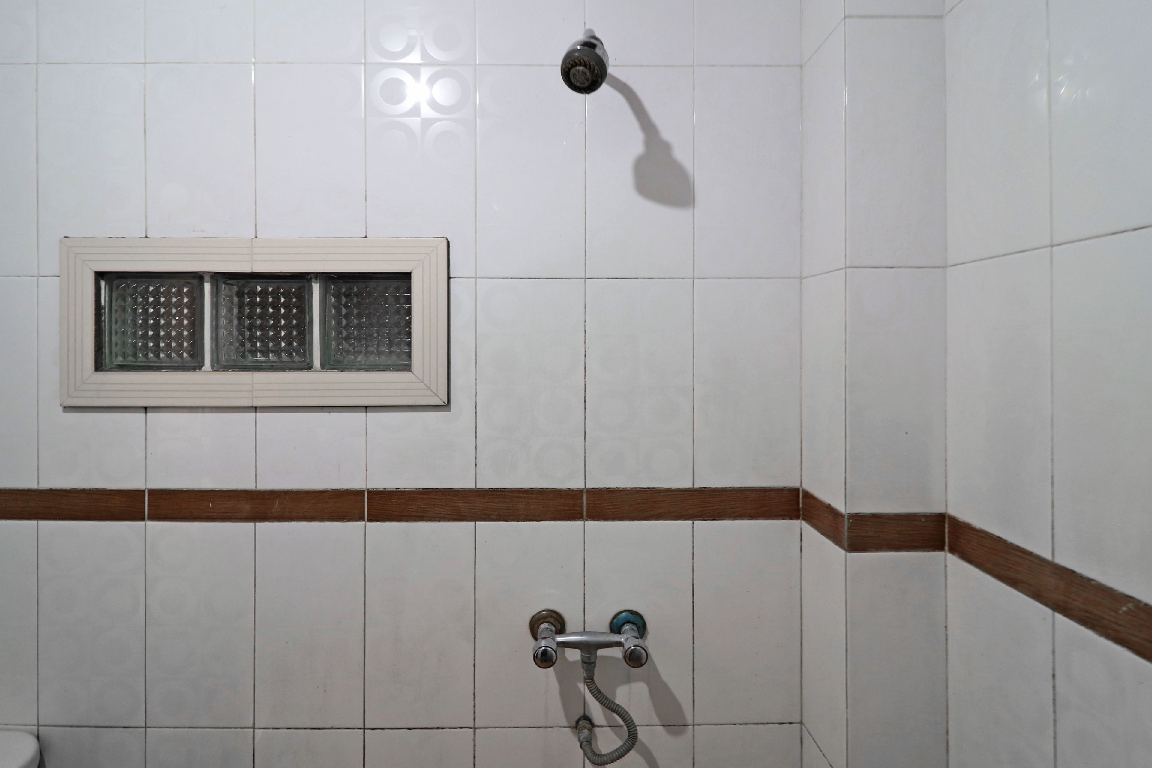 A bathroom with white tiled walls, a window with a brown frame, and a shower with a hose.