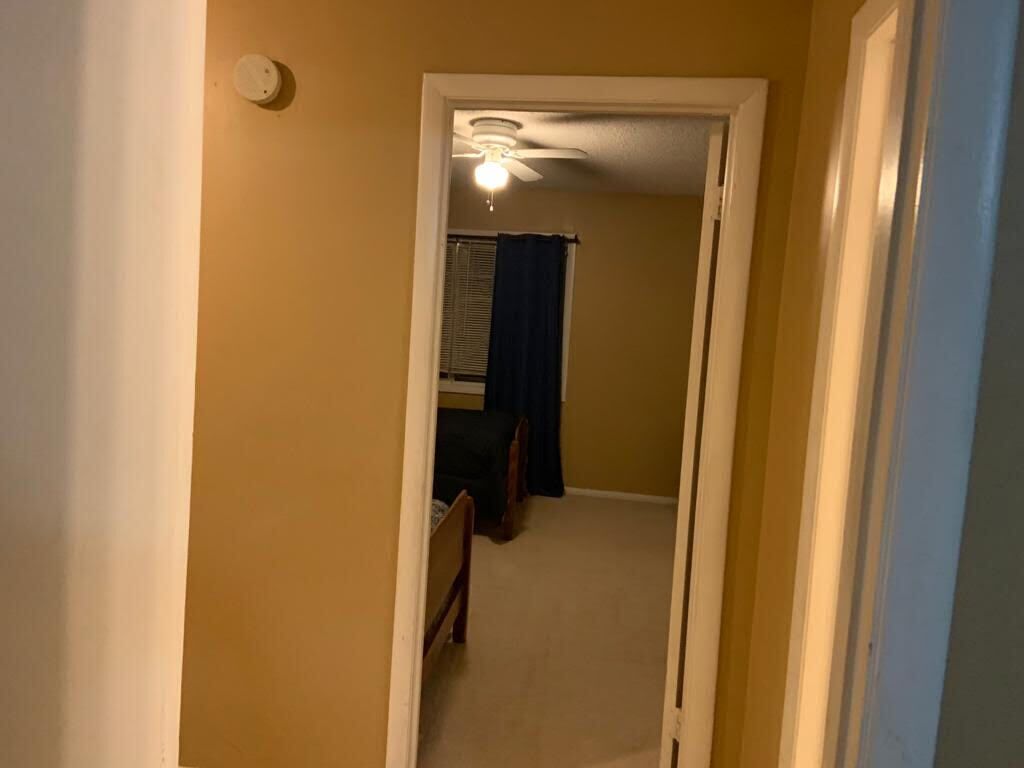 A narrow hallway with a ceiling fan and a door leading to a room with a bed and a window with curtains.
