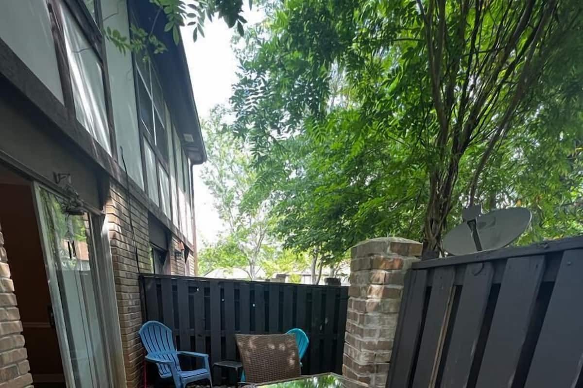 An outdoor patio area with a concrete floor, a brick wall, and a black metal fence. There are two wicker chairs and a blue plastic chair.