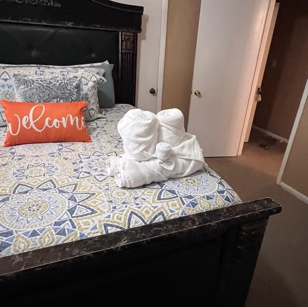 A bed with a patterned bedspread, a 'Welcome' pillow, and a stack of white towels.