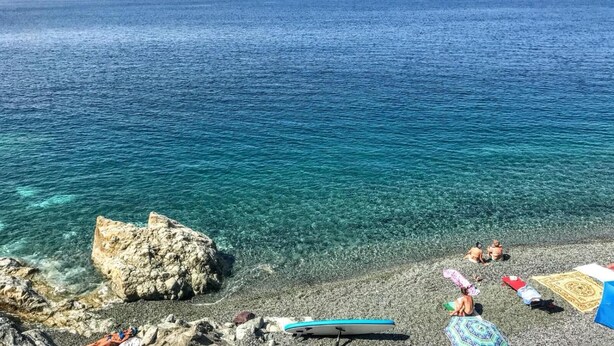Ein Strand mit klarem türkisblauem Wasser, Kieselsteinen und einer großen Felsformation.