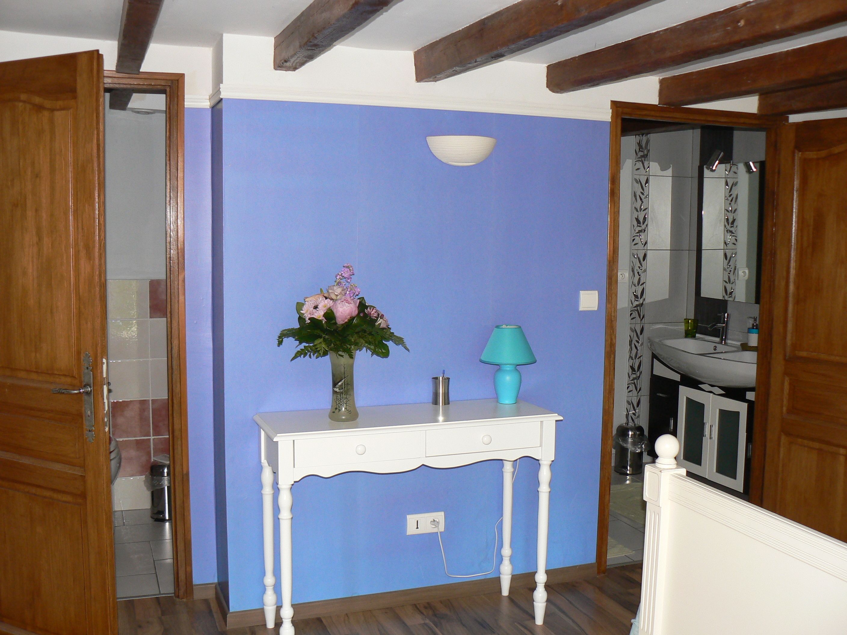 Een badkamer met een blauwe wand, houten balken, een wit wastafelmeubel en een vaas met bloemen.