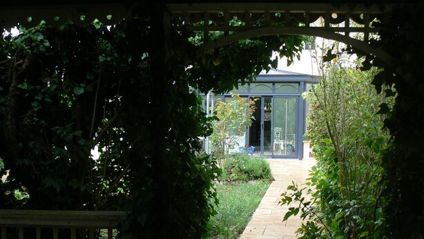 A garden pathway leading to a conservatory with a glass door, surrounded by lush greenery. - La Maison de l'étang (Sampigny)