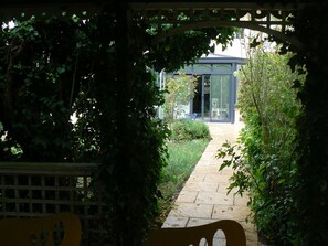 A garden pathway leading to a conservatory with a glass door, surrounded by lush greenery. - La Maison de l'étang (Sampigny)