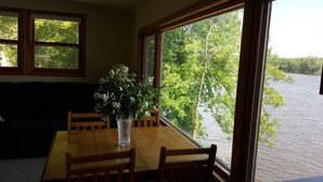 A dining area with a wooden table, chairs, and a vase of flowers. Large windows provide a view of a body of water and trees.