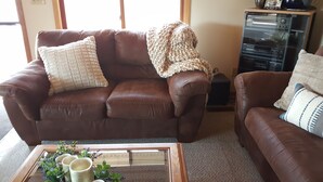 A brown leather sofa with a knitted throw blanket and a cushion. A small wooden coffee table with a glass top and decorative items is in front.