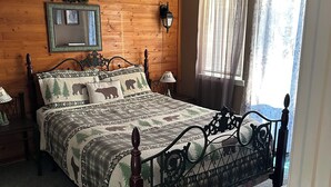 Un dormitorio con pared de madera, cama con colcha floral, mesita de noche, lámpara y ventana con cortinas.