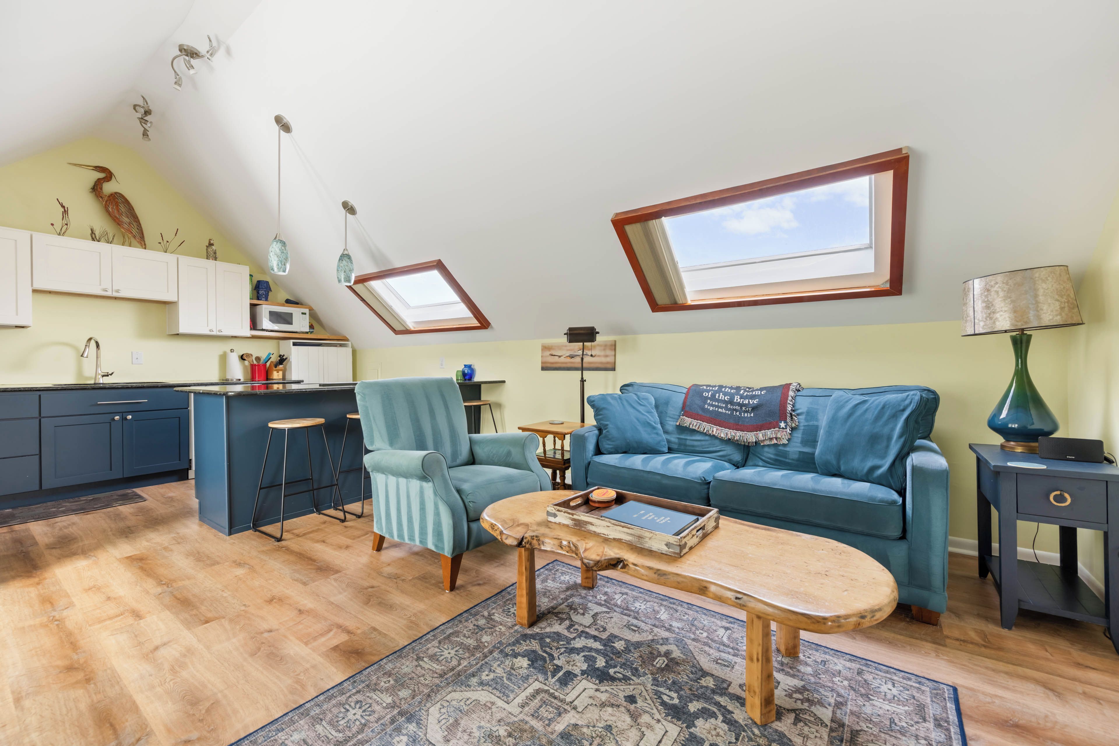 Ein modernes Wohnzimmer mit Sofa, Sessel, Couchtisch und einer Küchenzeile mit Schränken und einer Mikrowelle.