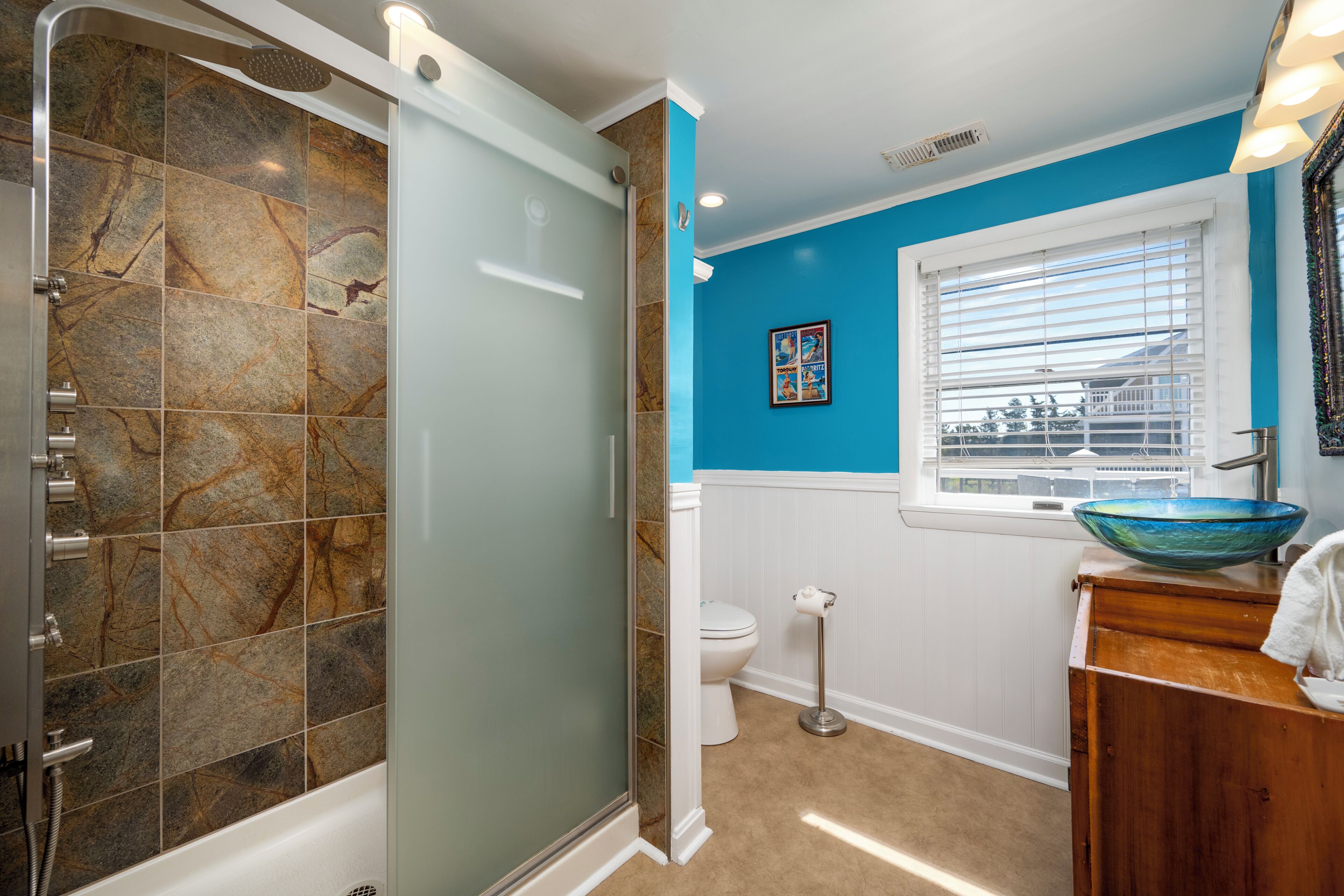 Une salle de bain avec une douche à parois de verre, des toilettes, un lavabo avec un récipient bleu et une fenêtre avec des stores.