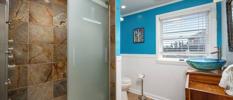 Une salle de bain avec une douche à parois de verre, des toilettes, un lavabo avec un récipient bleu et une fenêtre avec des stores.