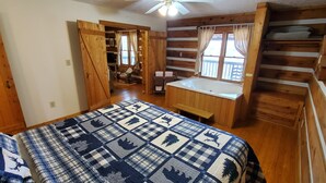 A wooden cabin bedroom with a large bed, a bench, a window with curtains, and a wooden dresser.