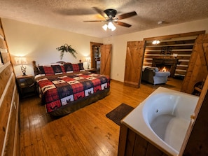 A bedroom with a large bed, a ceiling fan, a wooden floor, a fireplace, and a bathroom with a bathtub.