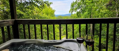 A hot tub with a glass of red wine and a candle on a wooden deck with a forest view.