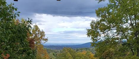 A view from a balcony with a wooden railing, overlooking a forested area with autumn foliage and distant mountains under a cloudy sky.