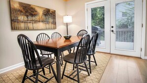 A dining area with a wooden table and black chairs, a painting on the wall, a floor lamp, and a sliding glass door with blinds.