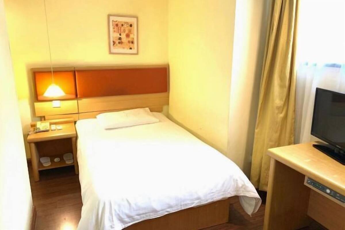 A small hotel room with a single bed, a bedside table with a lamp, a TV, and a picture on the wall.