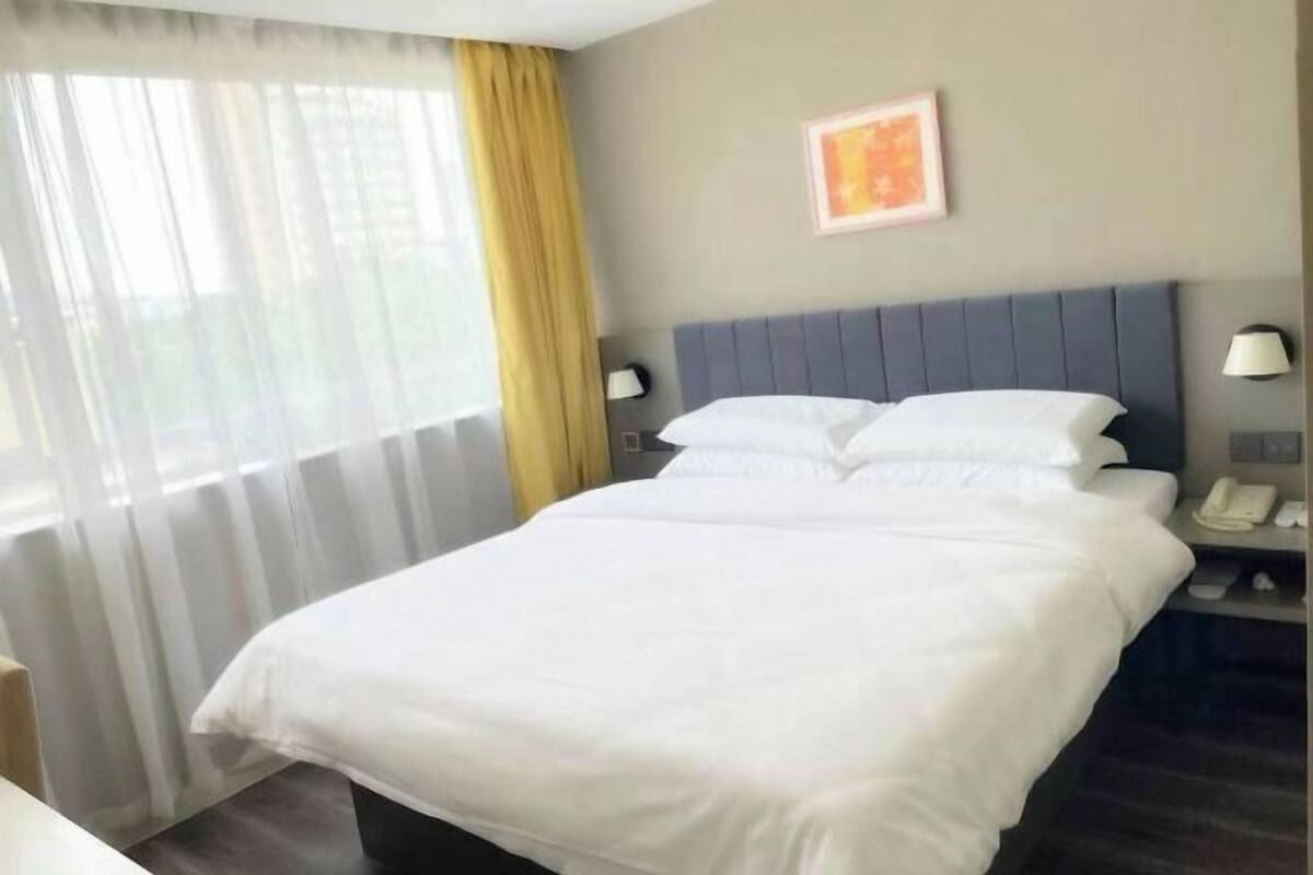 A hotel room with a bed, a nightstand with a phone, a window with curtains, and a wall-mounted picture.
