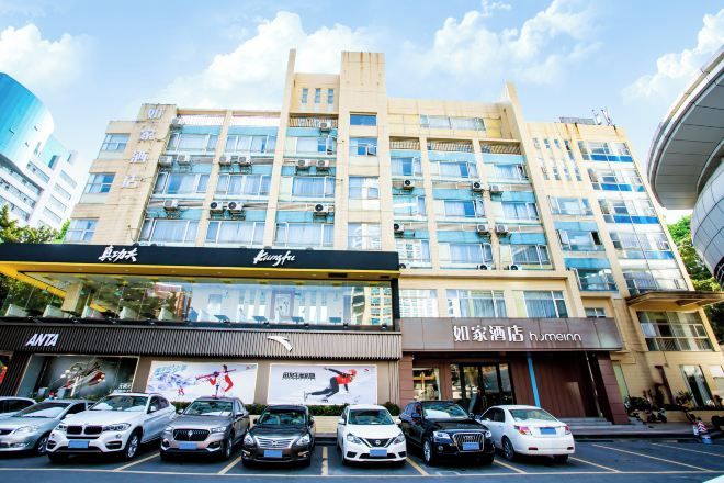 A multi-story hotel with a modern facade, several ground-level shops, and a parking lot with cars.