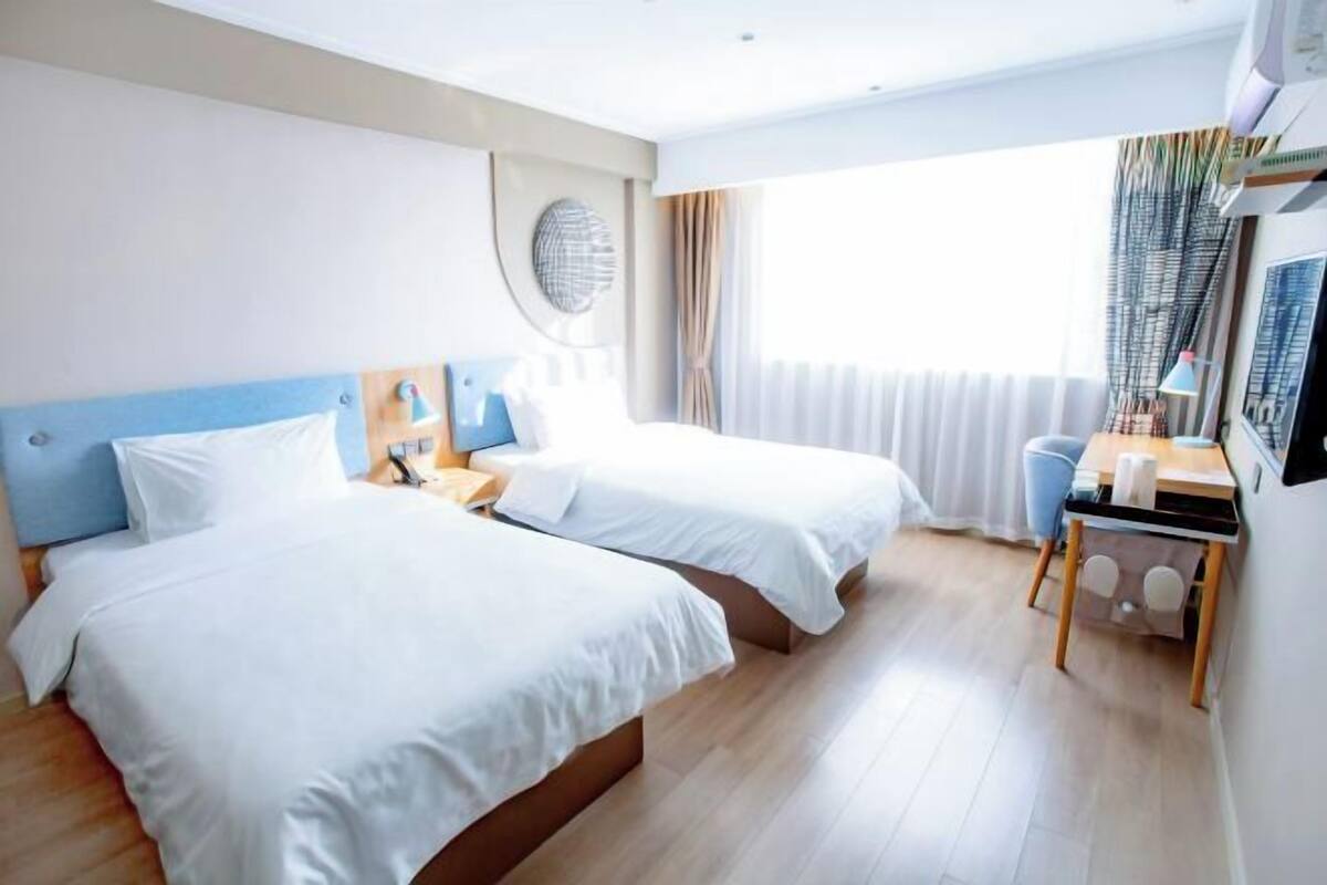 A hotel room with two beds, a desk, a chair, a lamp, and a window with curtains.