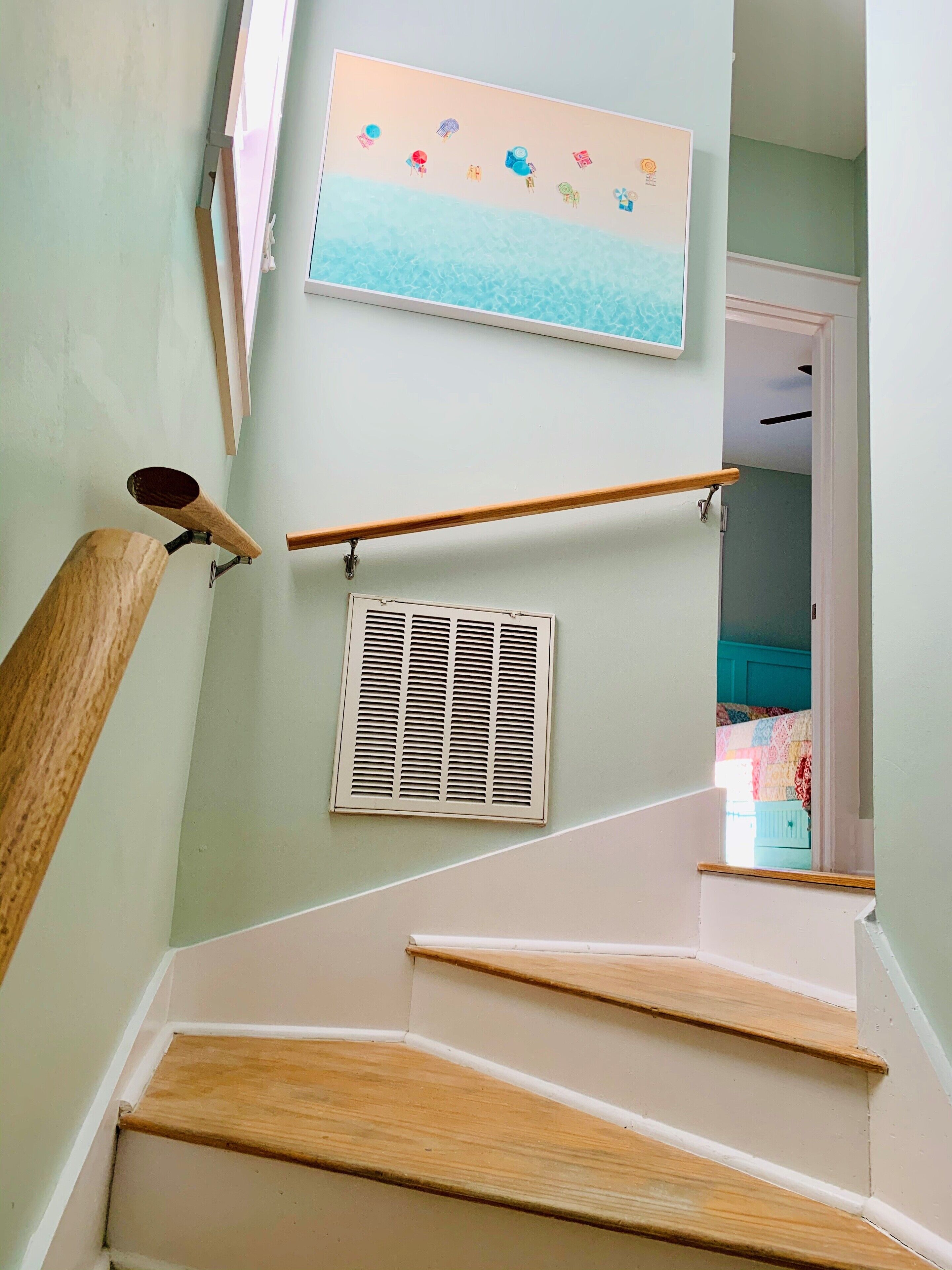 Eine Treppe mit Holzbalken und Handlauf, ein Wandlüfter und ein Strandmotiv über der Treppe.