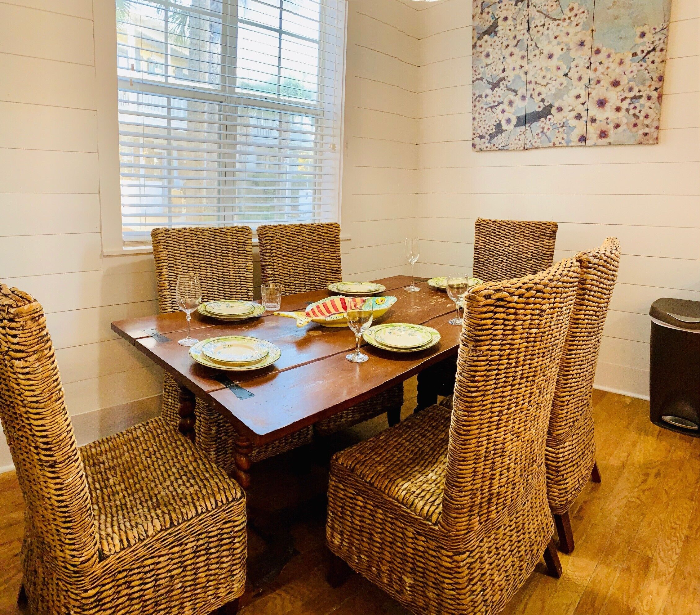 A dining area with a wooden table and wicker chairs, a window with blinds, and a painting on the wall.