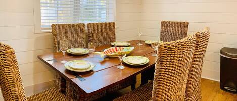 A dining area with a wooden table and wicker chairs, a window with blinds, and a painting on the wall.
