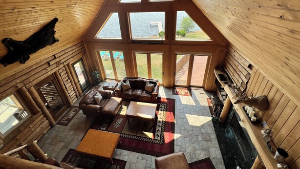 L’intérieur d’une cabane en rondins, avec un plafond haut, des murs en bois, une cheminée, un canapé en cuir et de grandes fenêtres donnant sur un lac.