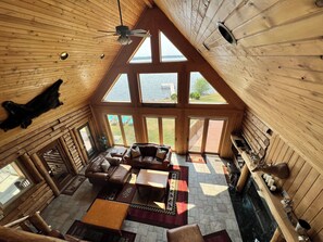 A log cabin interior with a high ceiling, wooden walls, a fireplace, a leather sofa, and large windows overlooking a lake. - Log Cabin w/indoor pool on Lake, 30 Min from Wis Dells! 8 min. from Sand Valley (Arkdale)