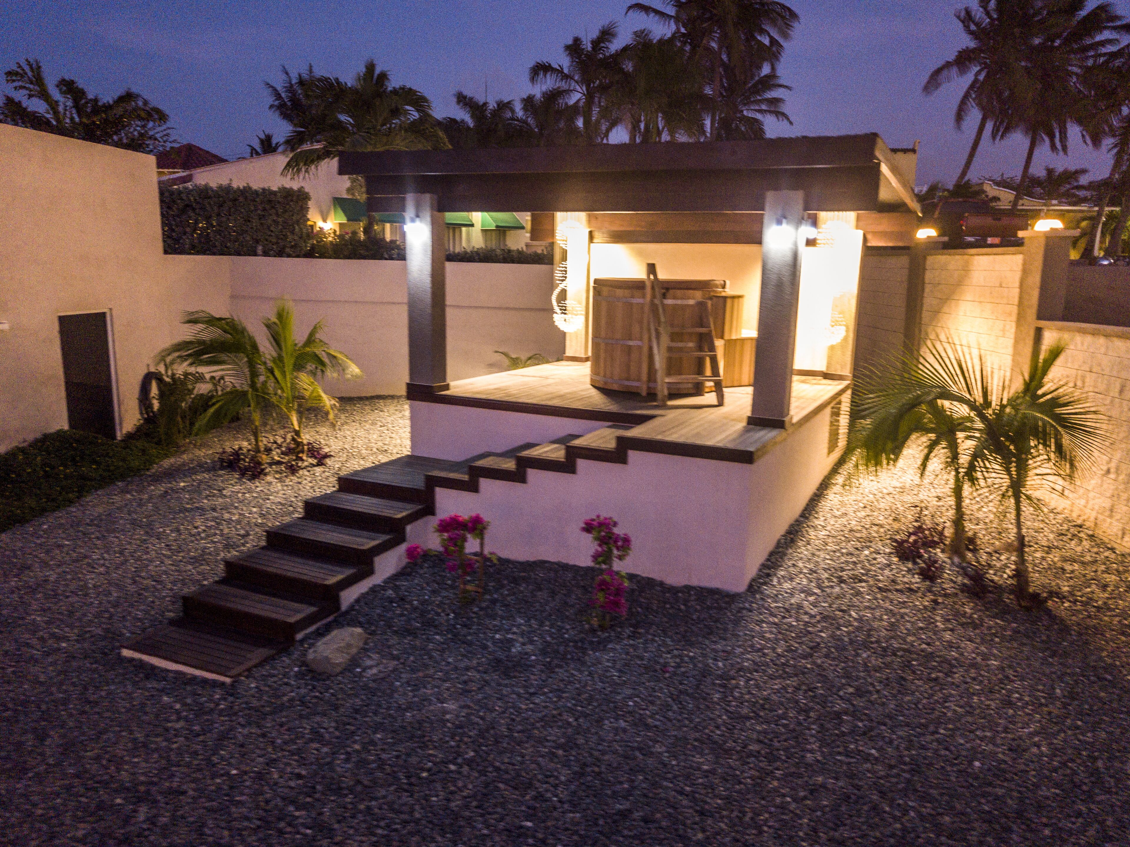 A modern outdoor patio with a wooden staircase leading to a covered seating area, surrounded by gravel and small plants.