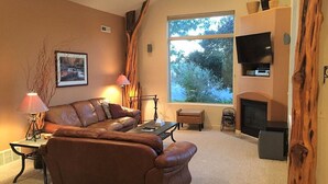 A living room with a brown leather sofa, a coffee table, a fireplace, a television on a shelf, and a large window with a view of trees. - Moab Rim Village Oasis (Moab)