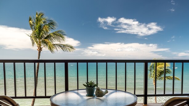 Mesa redonda con dos sillas en un balcón con vista a una playa con palmeras y cielo despejado.