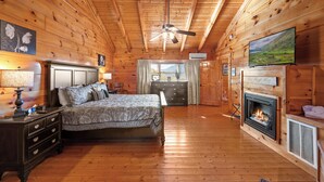 Ein Schlafzimmer in einer Blockhütte mit Bett, Kommode, Fernseher, Kamin und Deckenventilator.