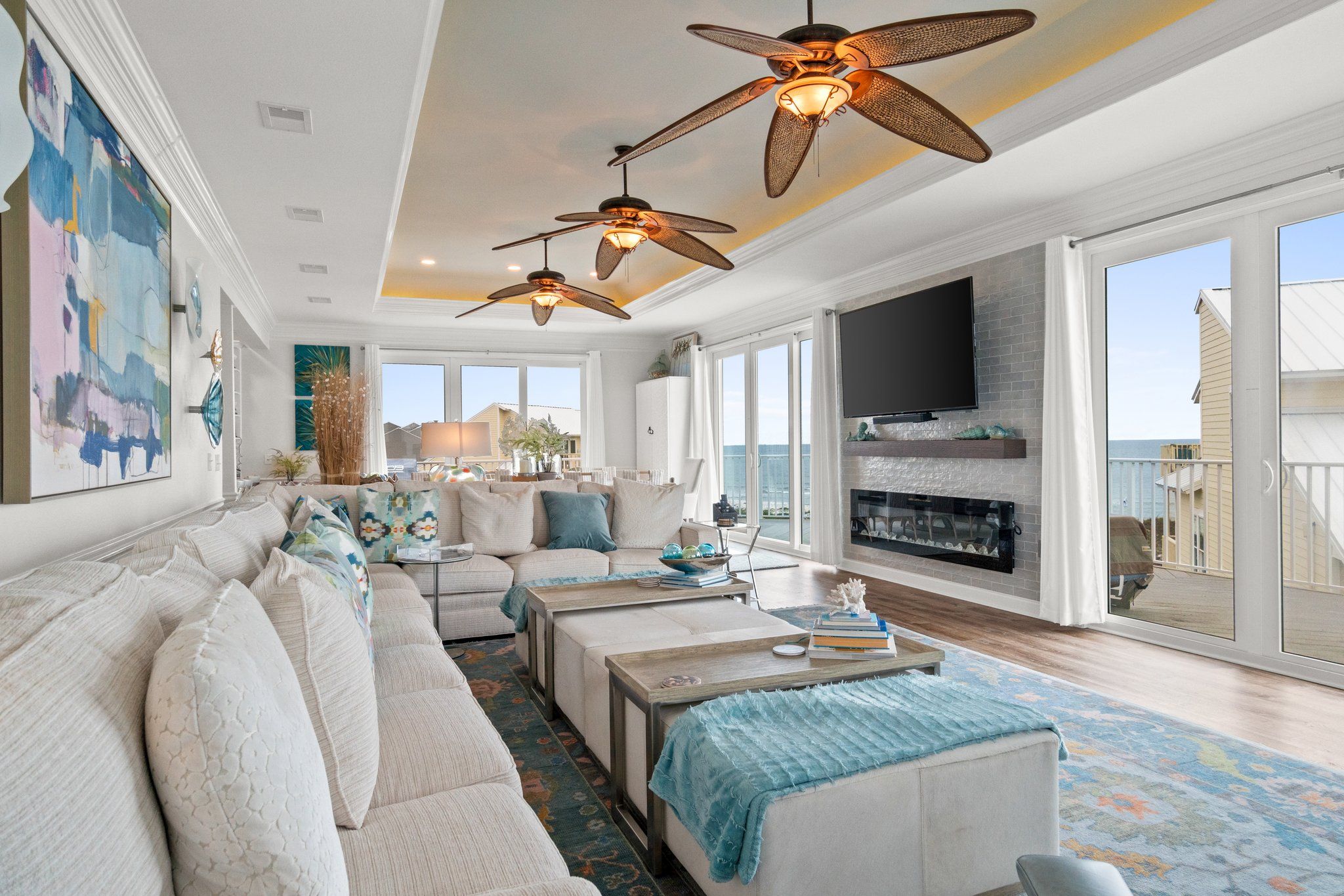 A spacious living room with a large sofa, two coffee tables, and a fireplace. There are ceiling fans, a TV, and a large window offering a view of the ocean.