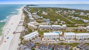 An aerial view of a coastal resort with beachfront properties, a swimming pool, and a boardwalk along the beach.