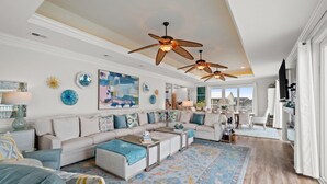 A spacious living room with a high ceiling, ceiling fans, a large sofa set, a coffee table, and a patterned rug.