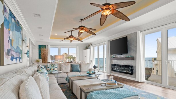 A spacious living room with a large sofa, two coffee tables, and a fireplace. There are ceiling fans, a TV, and a large window offering a view of the ocean.