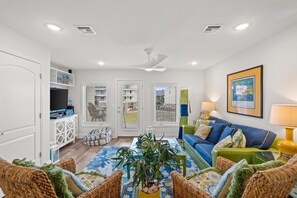 A living room with a blue sofa, green armchair, and a coffee table. There is a white cabinet with decorative items, a television, and a ceiling fan.