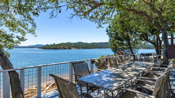 An outdoor dining area with a view of a lake and hills, furnished with metal chairs and tables, and a barbecue grill.