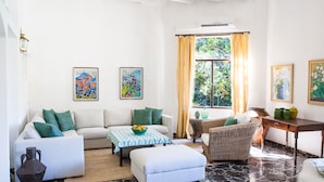 A living room with a sofa, ottoman, coffee table, and wicker chair. There are paintings on the walls and a window with curtains. - Stunning Villa and Garden w/ Pool and Sweeping Views in the Heart of Fornalutx (Fornalutx)