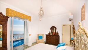 Un dormitorio con una cama grande, un tocador, un espejo y vista al mar a través de una ventana abierta.