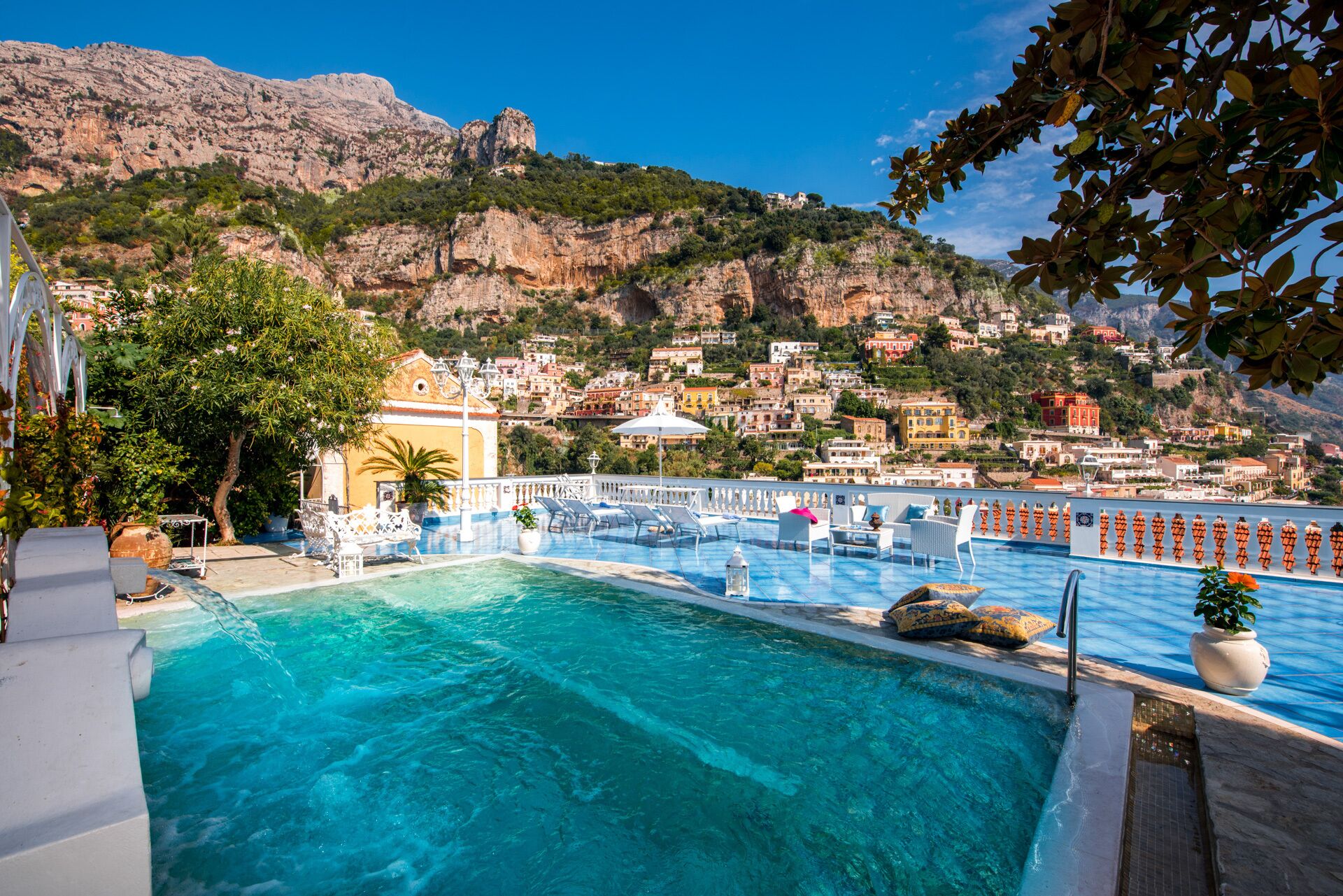 A pool area with clear blue water, white lounge chairs, and a view of a hillside town with buildings.