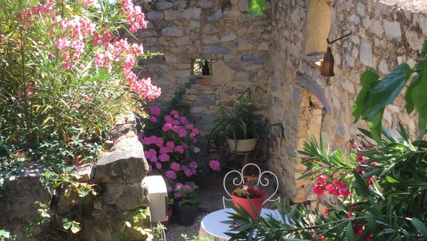 Un patio con paredes de piedra, una mesa y sillas, rodeado de plantas en macetas y arbustos florecientes.