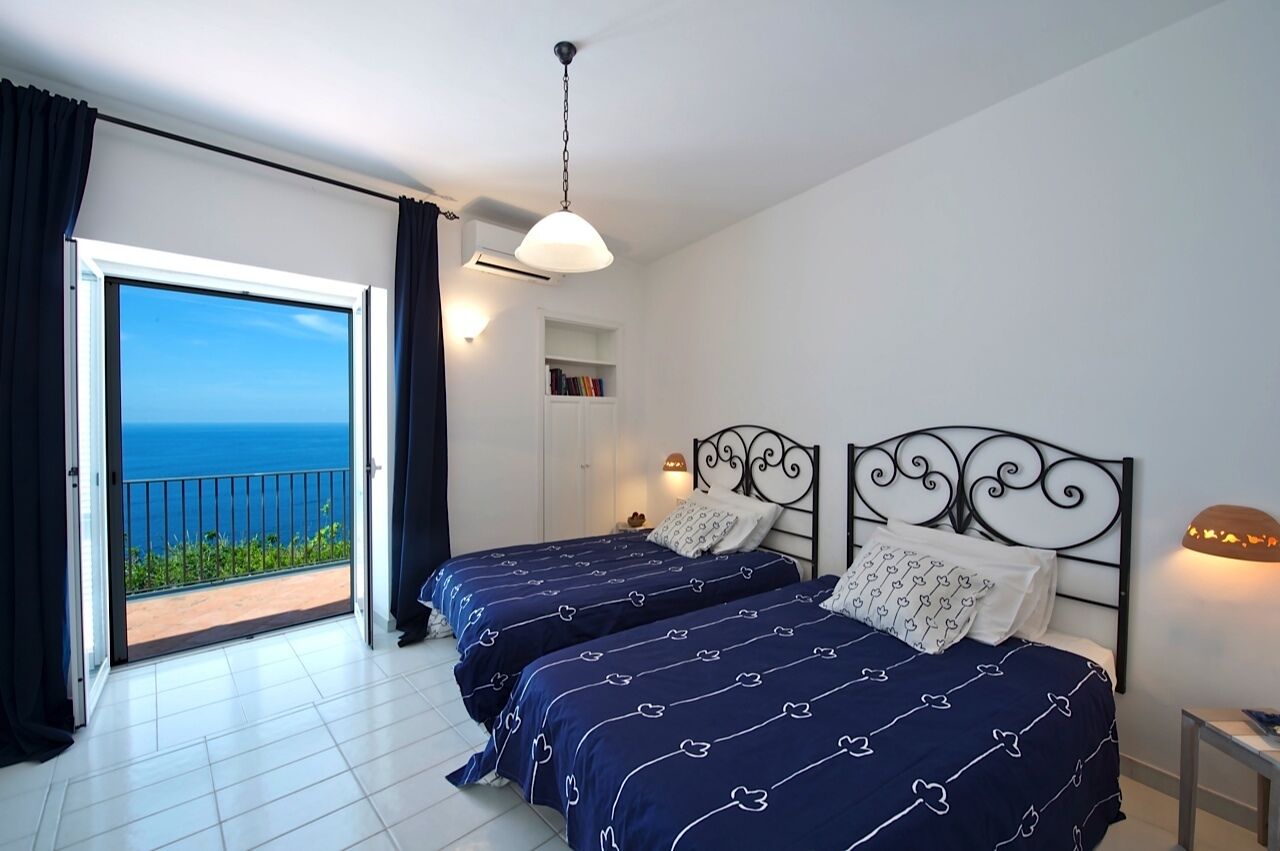 A bedroom with two beds, a balcony with a view of the sea, and a ceiling light.