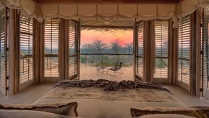 A room with a large bed, a view of the sunset through open windows, and a balcony with a chair. - Ruaha Dream Villa (Ruaha National Park)