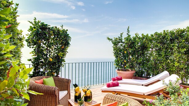 A balcony with wicker furniture, a table with drinks, and potted plants. - Villa Luce (Praiano)