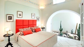 A bedroom with a red headboard, a bed with white and red bedding, framed pictures on the wall, and a cactus garden in the corner. - Villa Luce (Praiano)