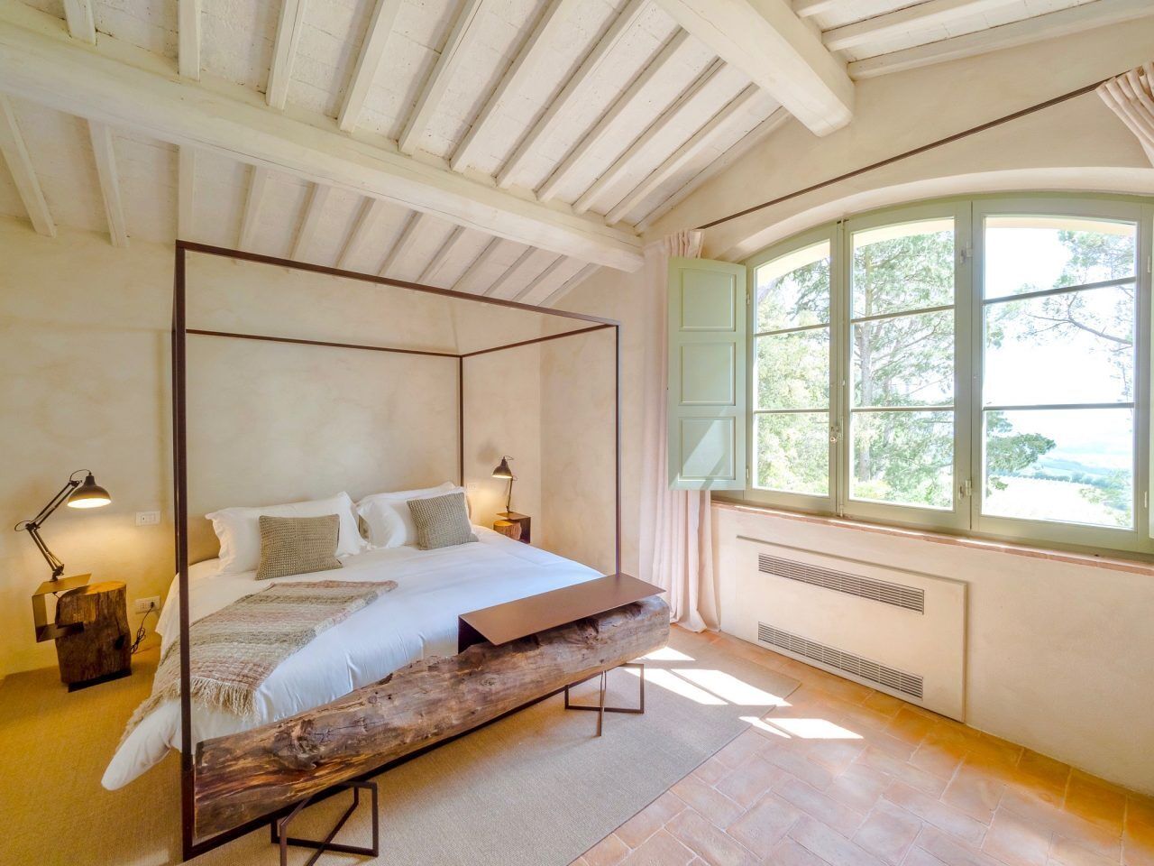 A bedroom with a four-poster bed, a window with curtains, a nightstand, and a lamp.