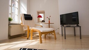 A living room with a wooden chair, a small table with a lamp, a TV on a stand, and a window with a view of buildings. - Haus Weihertor (Traben-Trarbach)