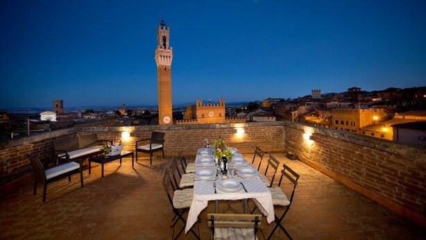 A rooftop dining area with a table set for six, overlooking a historic town with a prominent tower. - Torre del Campo (Siena)