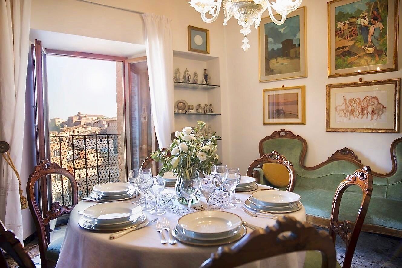 A dining room with a table set for six, a vase of flowers, and a view of a cityscape through an open window.
